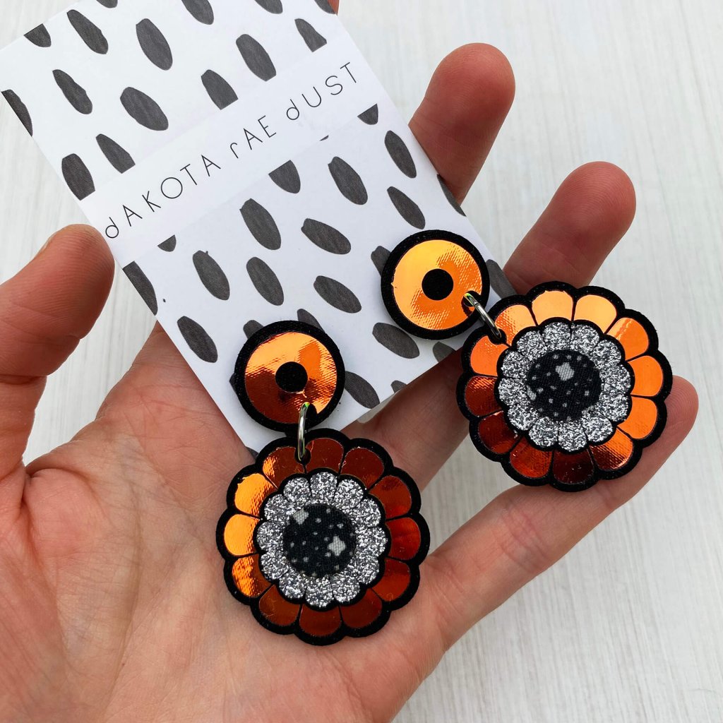 A pair of iridescent floral motif earrings mounted on a black and white patterned, dakota rae dust branded card are held in an open hand.