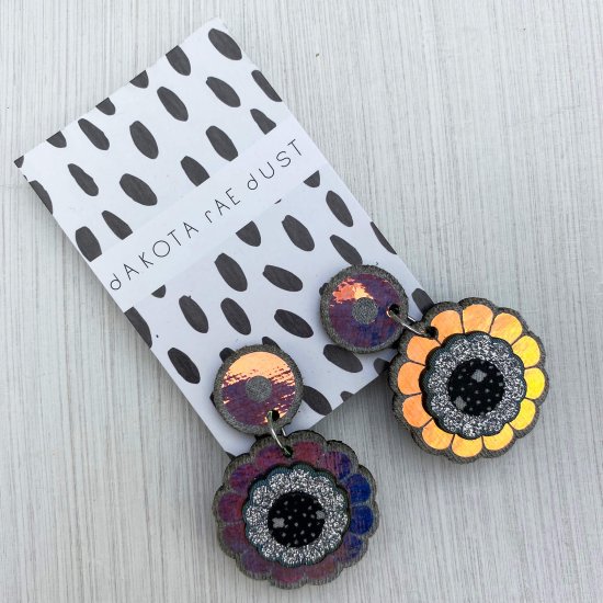 A pair of mini iridescent flower earrings mounted on a black and white patterned, dakota rae dust branded card.