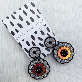 A pair of mini glittery floral earrings mounted on a black and white patterned, dakota rae dust branded card.