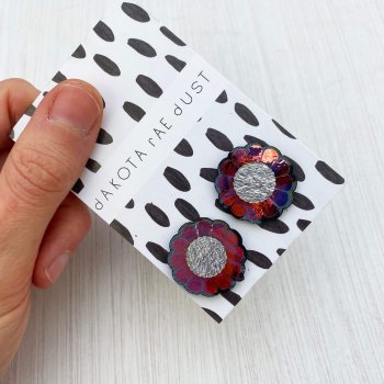 A pair of festive flower studs mounted on a black and white patterned, dakota rae dust branded card are held by a just visible thumb and forefinger