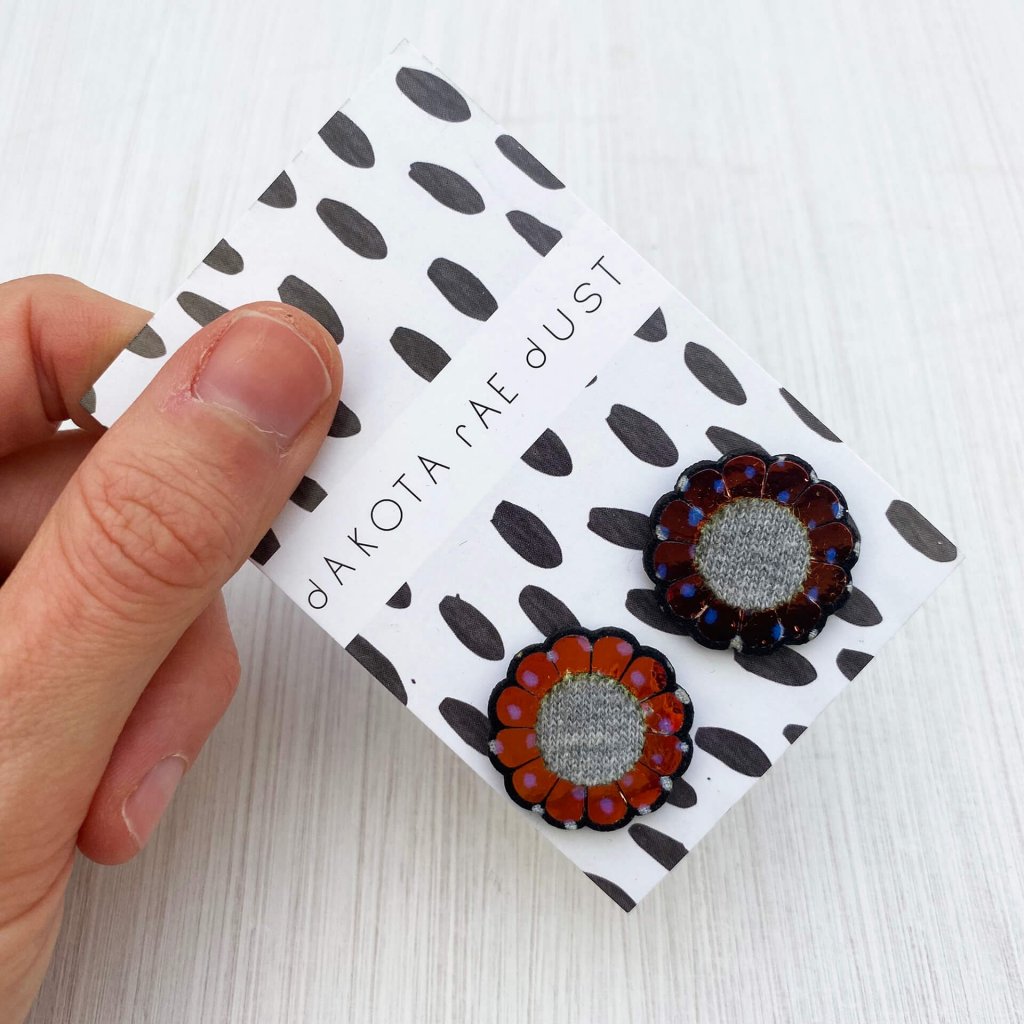 A pair of iridescent flower studs mounted on a black and white patterned, dakota rae dust branded card are held by a just visible thumb and forefinger