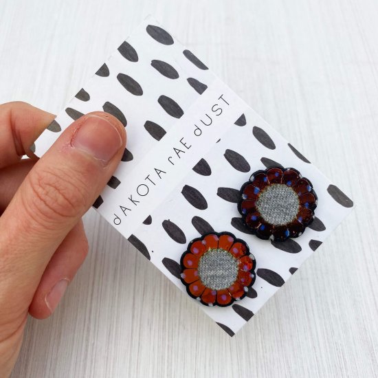 A pair of iridescent flower studs mounted on a black and white patterned, dakota rae dust branded card are held by a just visible thumb and forefinger