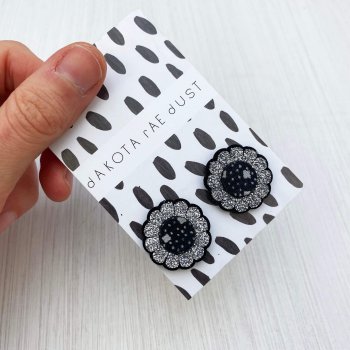 A pair of glittery floral stud earrings mounted on a black and white patterned, dakota rae dust branded card are held by a just visible thumb and forefinger