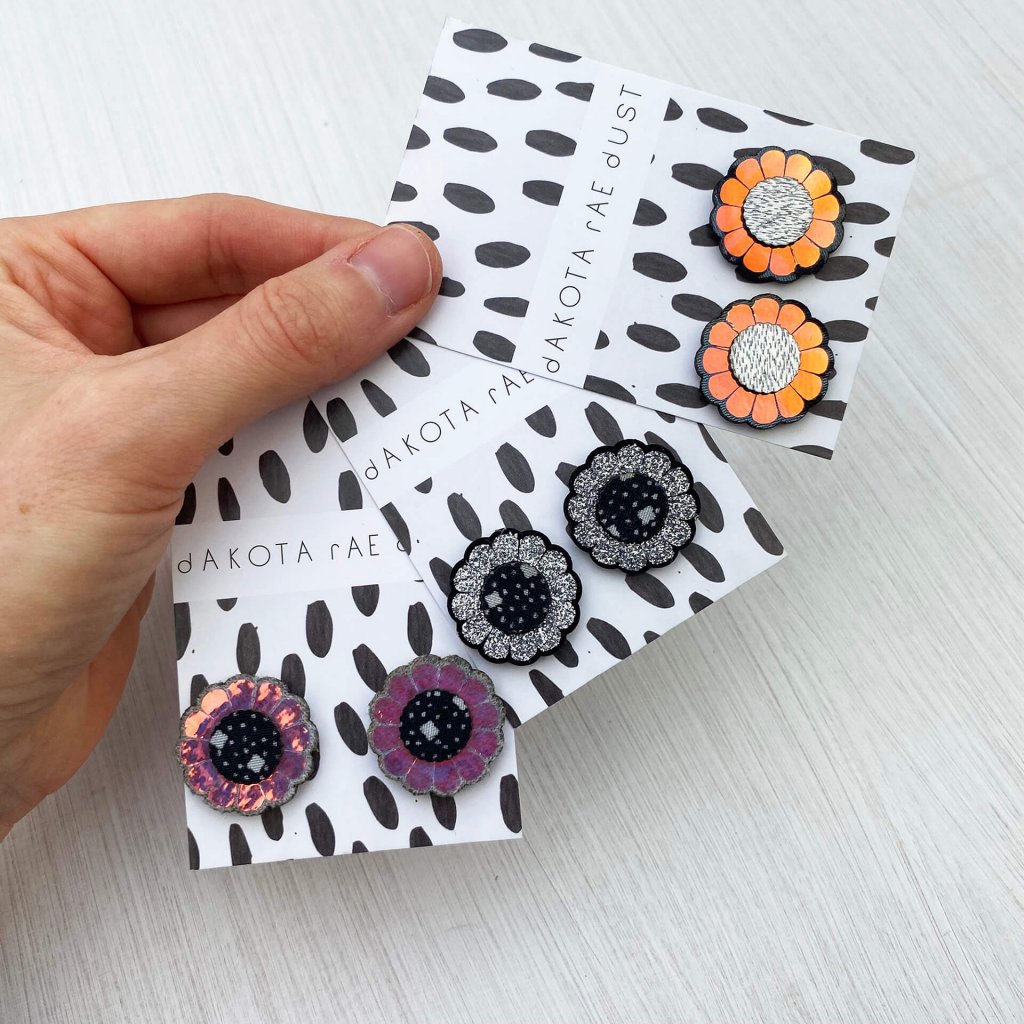 A woman's hand holds three pairs of sparkly floral studs mounted on black and white patterned, dakota rae dust branded cards