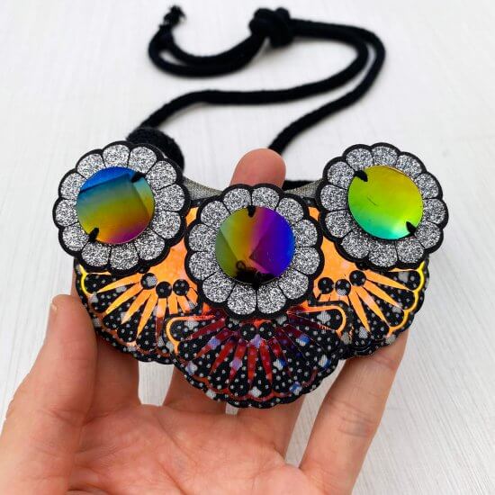 An ornate and iridescent bib necklace with a row of three flower motifs and petrol coloured middles is held up by a white hand.