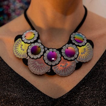 A moodily lit close up of a neck and chest area of someone wearing a wide V neck top, focusing on their black, silver glitter and grey oversize iridescent bib necklace