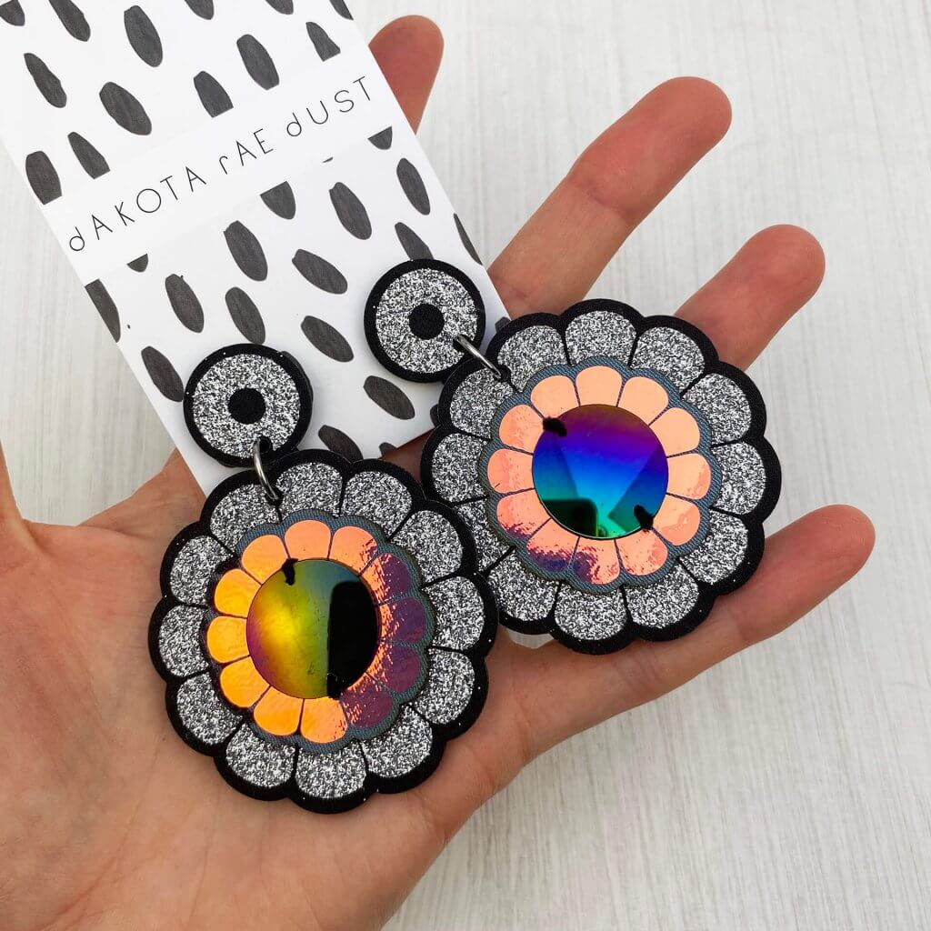 A pair of oversize flower motif earrings mounted on a black and white patterned, dakota rae dust branded card are held in an open hand.