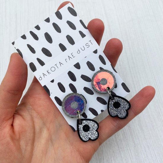 A pair of small dangly stud earrings mounted on a black and white patterned, dakota rae dust branded card are held by a just visible thumb and forefinger