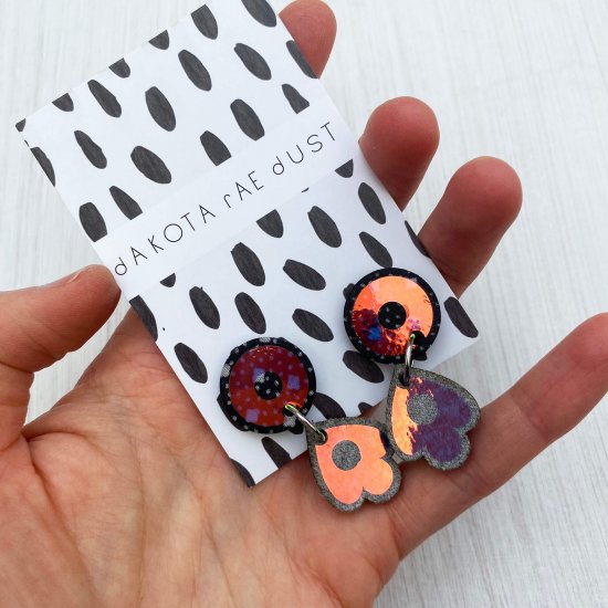 A pair of iridescent dangly studs mounted on a black and white patterned, dakota rae dust branded card are held by a just visible thumb and forefinger