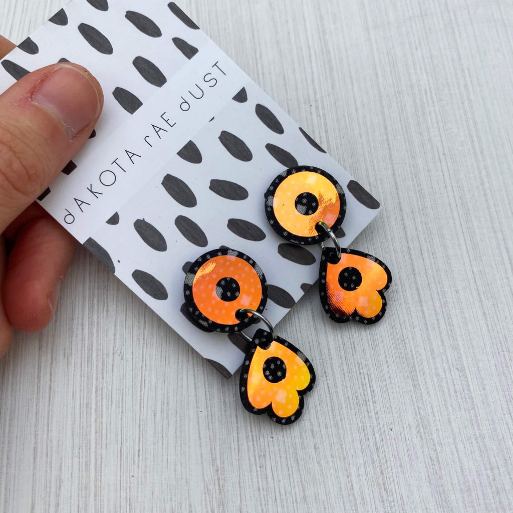 A pair of dainty dangly stud earrings mounted on a black and white patterned, dakota rae dust branded card are held by a just visible thumb and forefinger