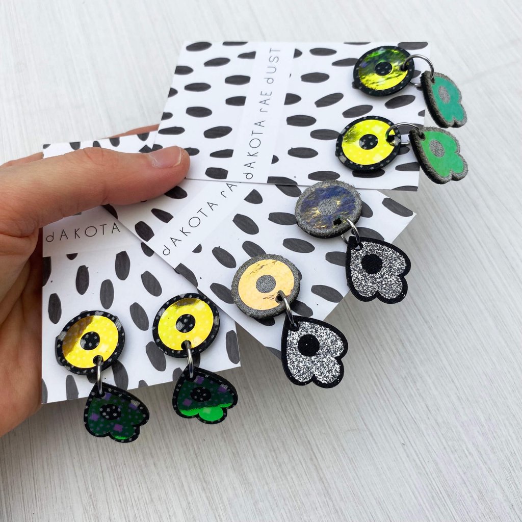 A woman's hand holds three pairs of dangly light reflecting studs mounted on black and white patterned, dakota rae dust branded cards