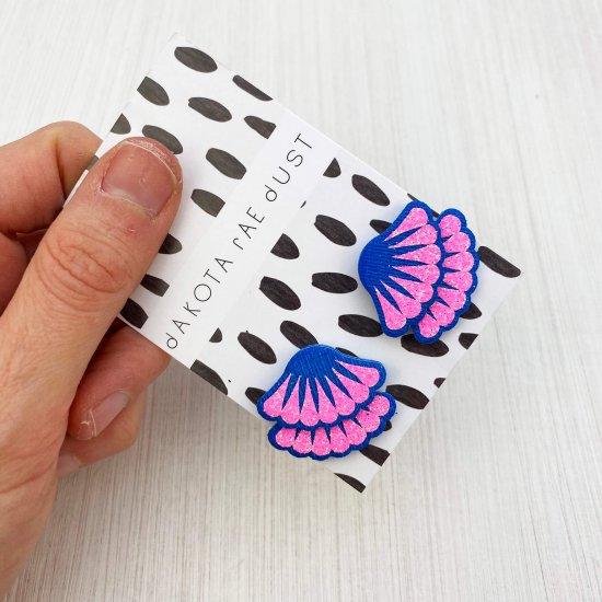 A pair of glittery pink and blue studs are mounted on a black and white patterned, dakota rae dust branded card and held in a woman's hand against an off white background