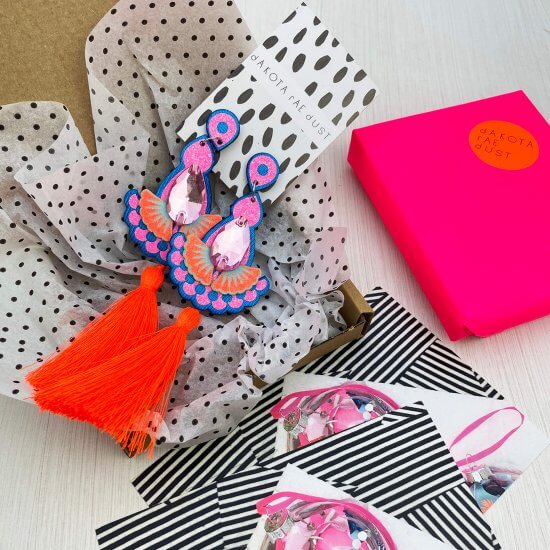 A partly unwrapped gift box filled with black and white polka dot patterned tissue paper and two pairs of neon orange statement tassel earrings sits next to a neon pink wrapped box and some colourful cards
