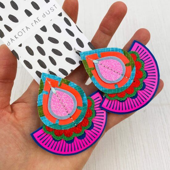 A pair a neon pink, blue and green oversize fan earrings on a black and white patterned dakota rae dust branded card, seen against a textured off white background