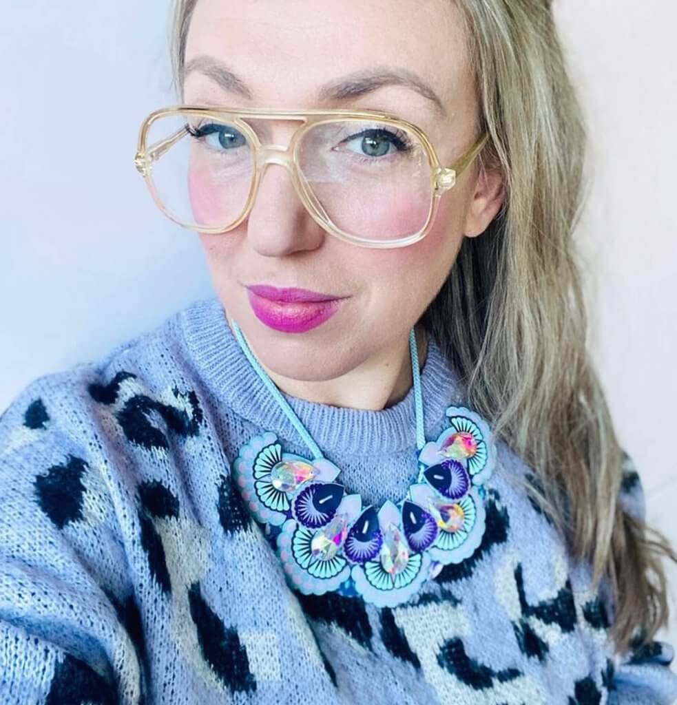 A blonde haired woman wearing large glasses and bright pink lipstick is showing 'how to style your statement bib necklace' by pairing hers with a lilac animal print woolly jumper.