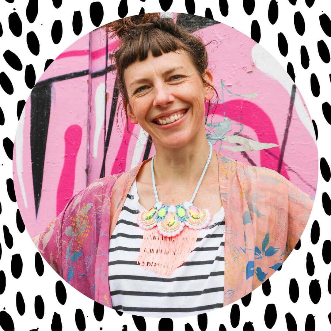 A white woman wearing a stripey t-shirt and statement bib necklace is smiling into the camera. Behind her is a pink graffiti covered wall. The round photo is framed by a block of white patterned with uneven, elongated polka dots.