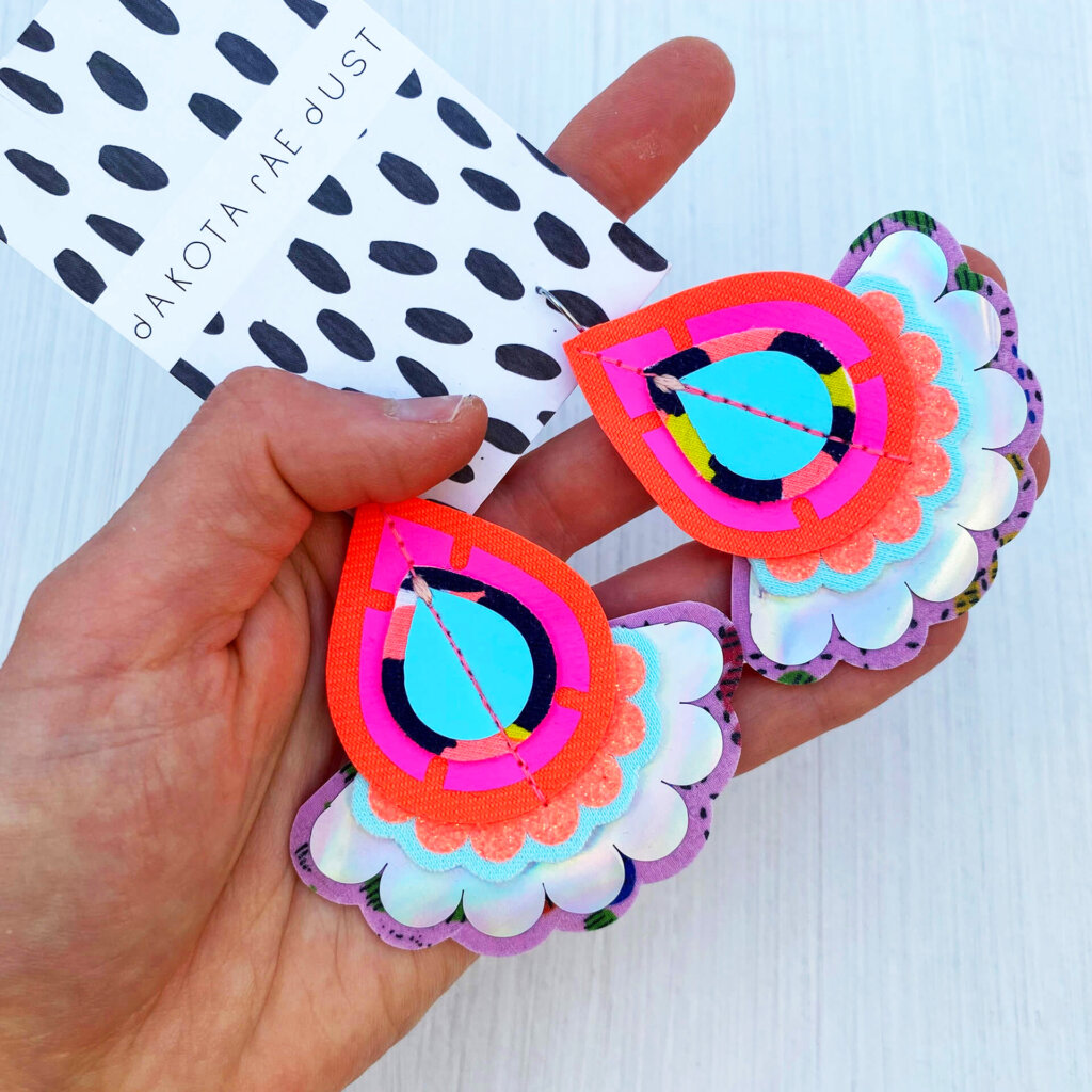 A pair of colourful oversize earrings with a holographic trim mounted on a black and white patterned, dakota rae dust branded card are lying on an off white background