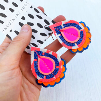 A pair of neon coral, orange and glittery blue recycled fabric earrings held in a open hand