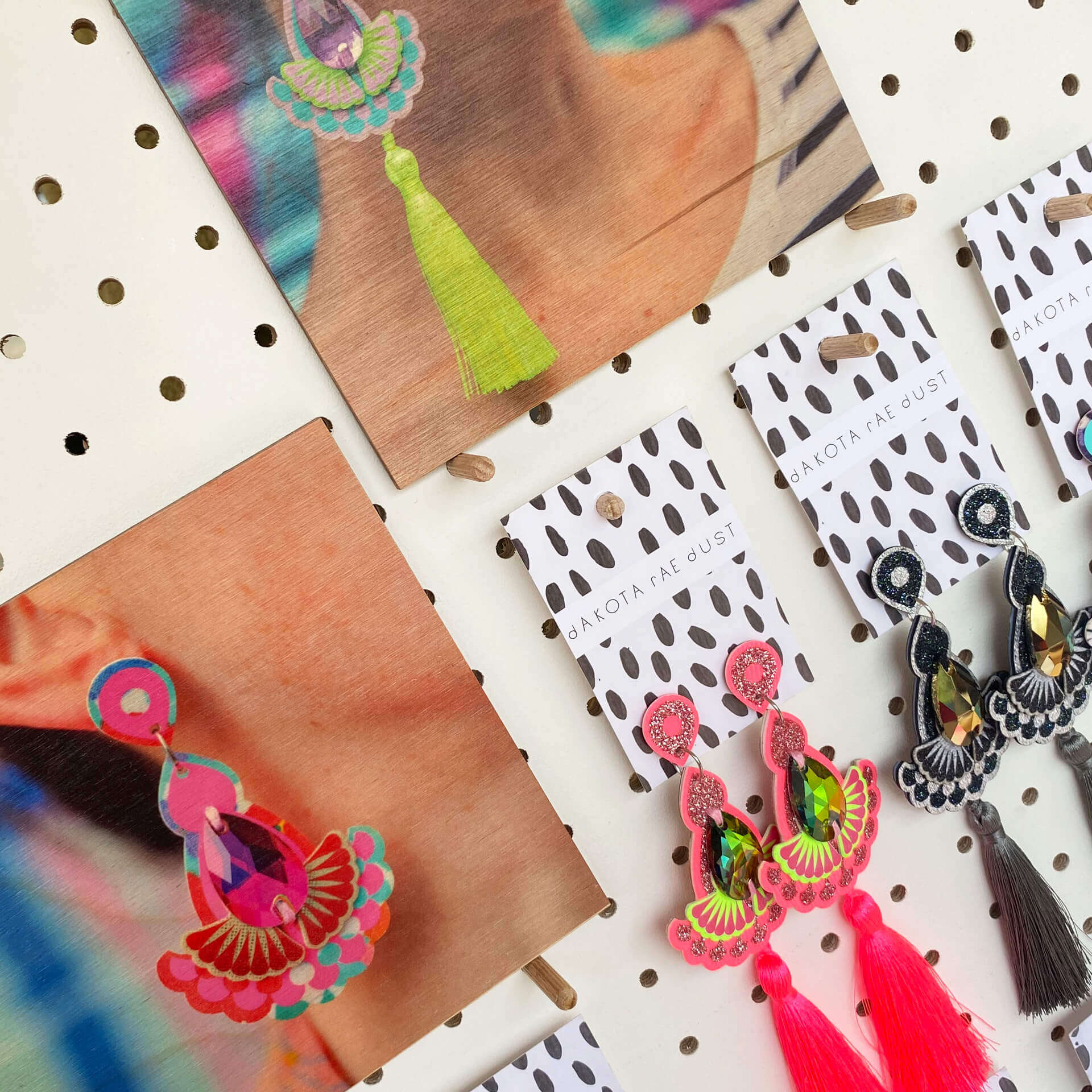 A close up of a selection of colourful tassel earrings and images of the earrings being modelled displayed on some white pegboard