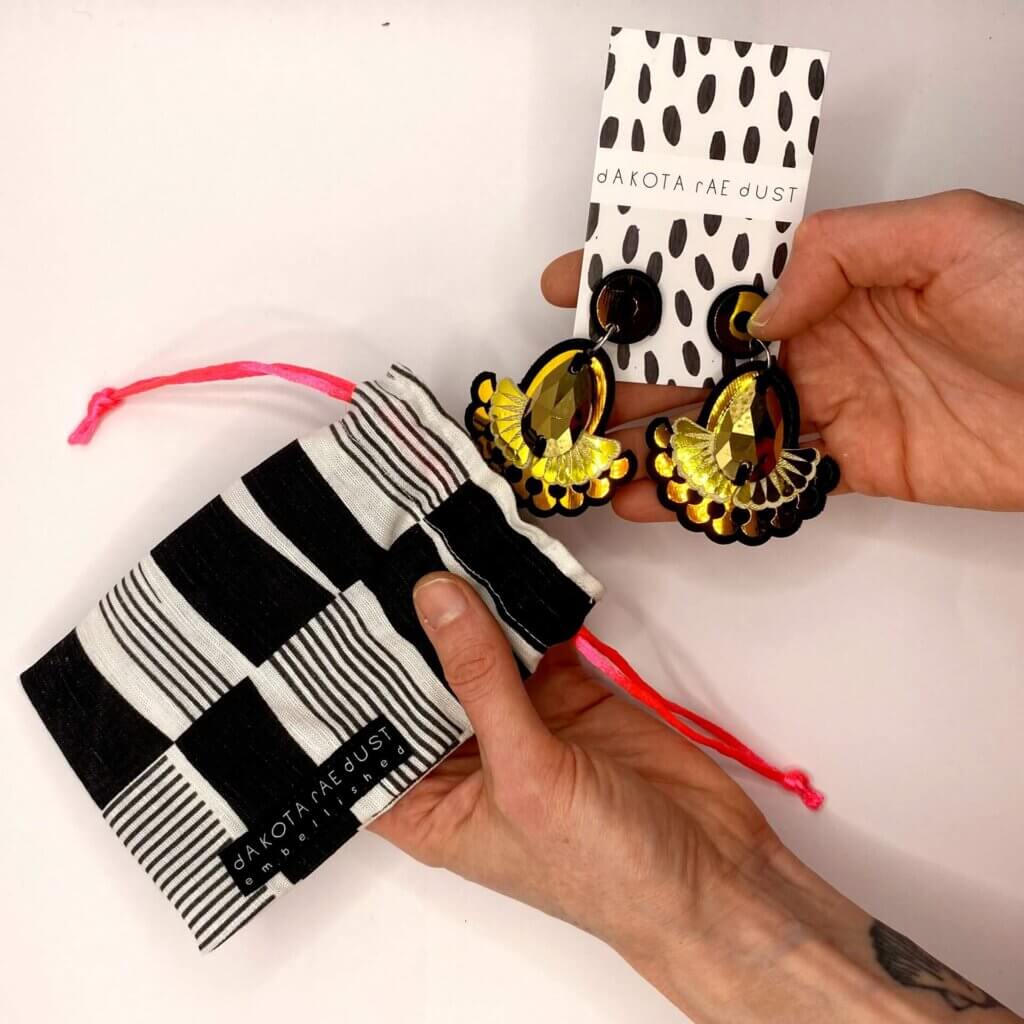 A pair ofblack and gold statement earrings mounted on a black and white patterned dakota rae dust branded card are being pulled from a black and white patterned drawstring jewellery bag with neon pink cords but a white pair of hands