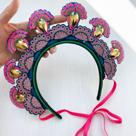 A colourful 7 jewel halo style headdress is held in a woman's hand against an off white background