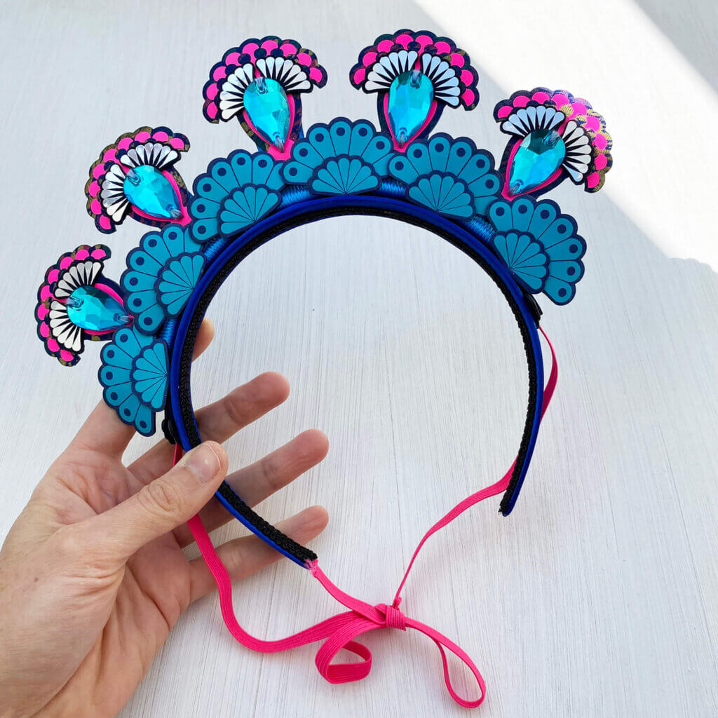 A colourful 5 jewel halo style headdress is held in a woman's hand against an off white background