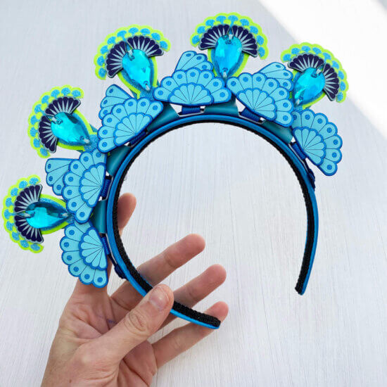 A bright blue and lime 5 jewel tiara style headdress is held in a woman's hand against an off white background