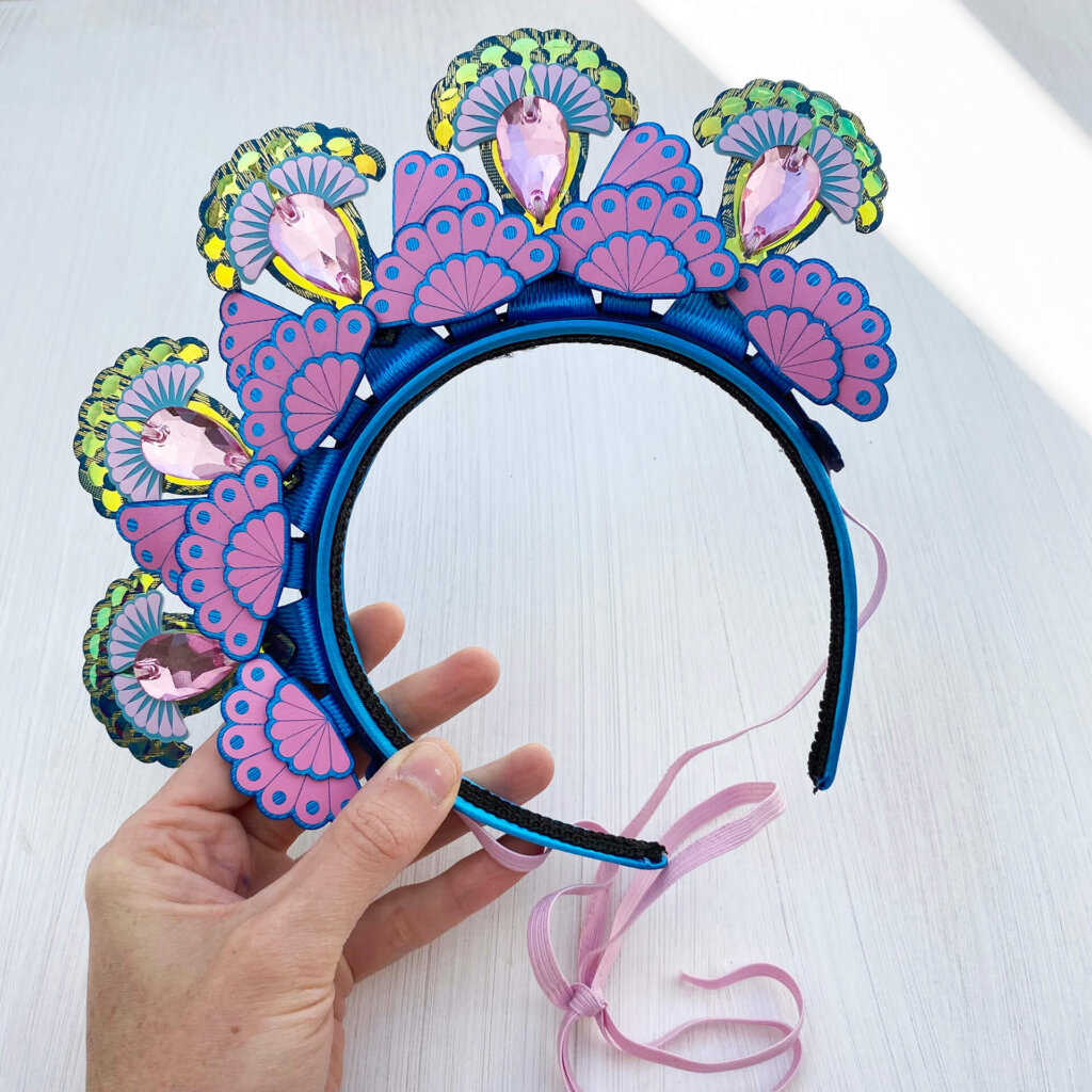 A blue and pink 5 jewel tiara style headdress is held in a woman's hand on an off white background