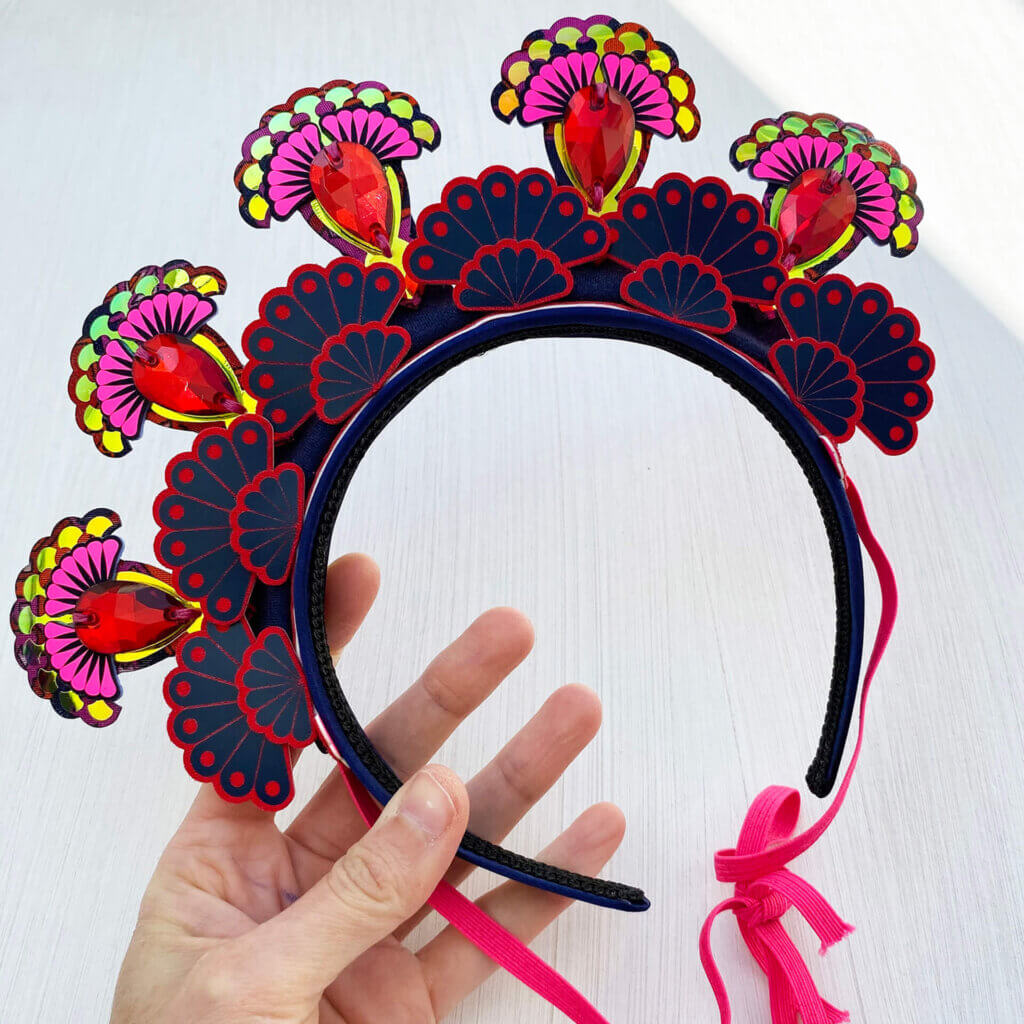 A colourful 5 jewel halo style, festival headdress is held in a woman's hand against an off white background