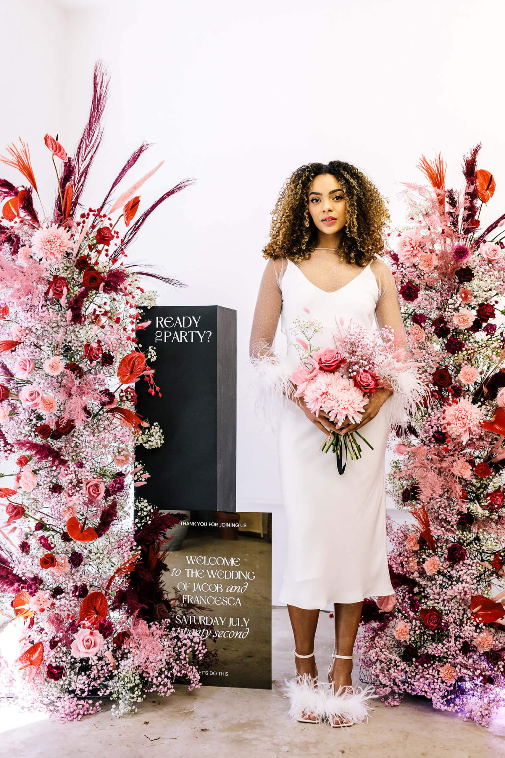 A young woman with curly brown hair wearing a wedding dress and holding a pink bouquet is starring into the camera. On either side of her is a tall sculptural, pink floral display. To her right is an engraved sign which reads 'ready to party?'