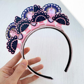 A pale pink and blue 3 jewel tiara headdress is held in a woman's hand on an off white background