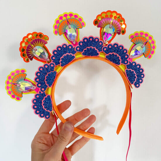 A colourful 5 jewel halo headdress is held in a woman's hand against an off white background