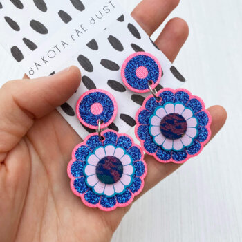 A pair of glittery blue and pink flower motif earrings are held in an open hand