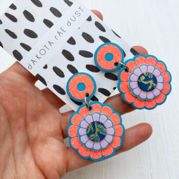 A pair of glittery orange and blue flower motif earrings are held in an open hand