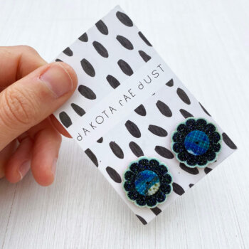 A pair of glittery floral studs mounted on a black and white patterned, dakota rae dust branded card are held by a just visible thumb and forefinger