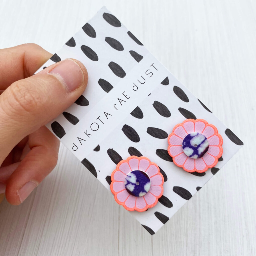 A pair of pretty pastel floral studs mounted on a black and white patterned, dakota rae dust branded card are held by a just visible thumb and forefinger