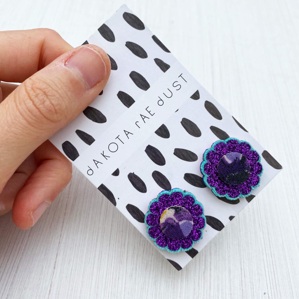 A pair of purple glittery floral studs mounted on a black and white patterned, dakota rae dust branded card are held by a just visible thumb and forefinger