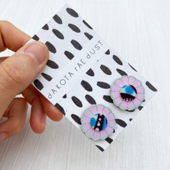 A pair of pretty pastel flower studs mounted on a black and white patterned, dakota rae dust branded card are held by a just visible thumb and forefinger