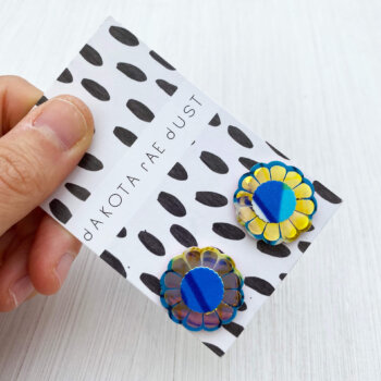 A pair of shiny gold floral studs mounted on a black and white patterned, dakota rae dust branded card are held by a just visible thumb and forefinger