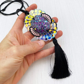 A custom cast giant jewel pendant necklace with a silky black tassel is held in an open hand on an off white background