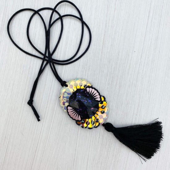 A custom cast giant jewel pendant necklace with a silky black tassel is lying on an off white background