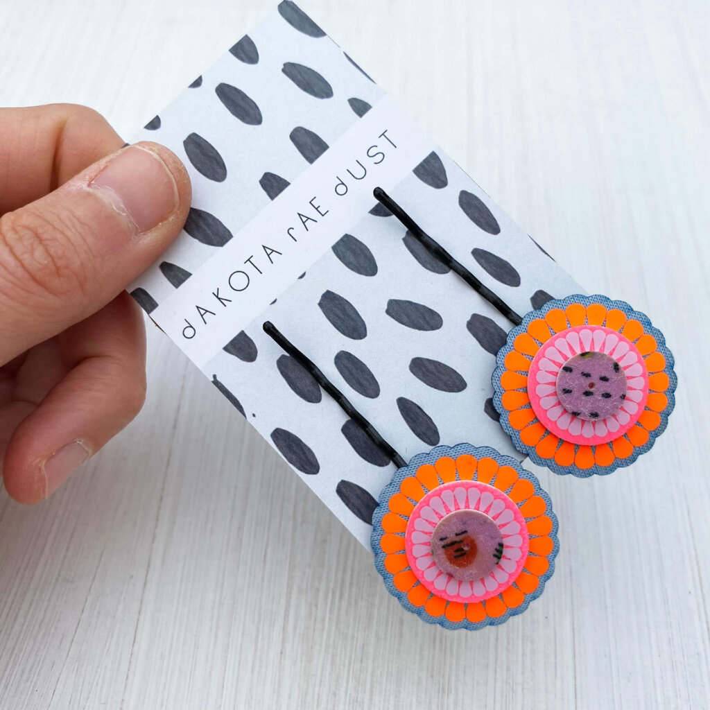 Fluorescent orange , light blue and lilac colourful hair pins on a black and white patterned backing card, held between the thumb and forefinger of a just visible hand
