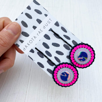 Fluorescent pink and navy colourful hair pins on a black and white patterned backing card, held between the thumb and forefinger of a just visible hand