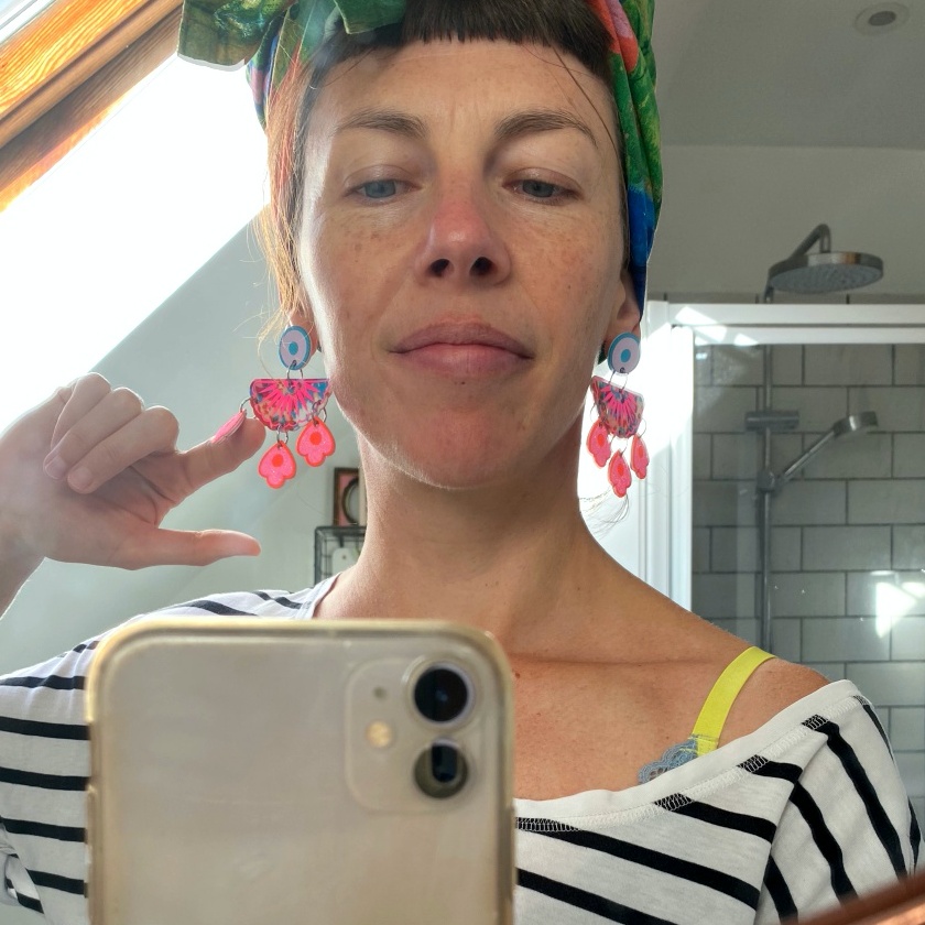 A woman wearing a stripey t-shirt, colourful statement earrings and a green head wrap is taking a selfie in a bathroom mirror