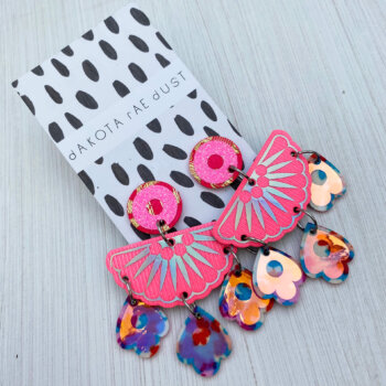 A pair of iridescent pink jangly droplet earrings mounted on an a black and white patterned dakota rae dust branded card are lying an off white background