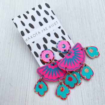 A pair of bright pink and turquoise jangly droplet earrings mounted on an a black and white patterned dakota rae dust branded card are lying on an off white background