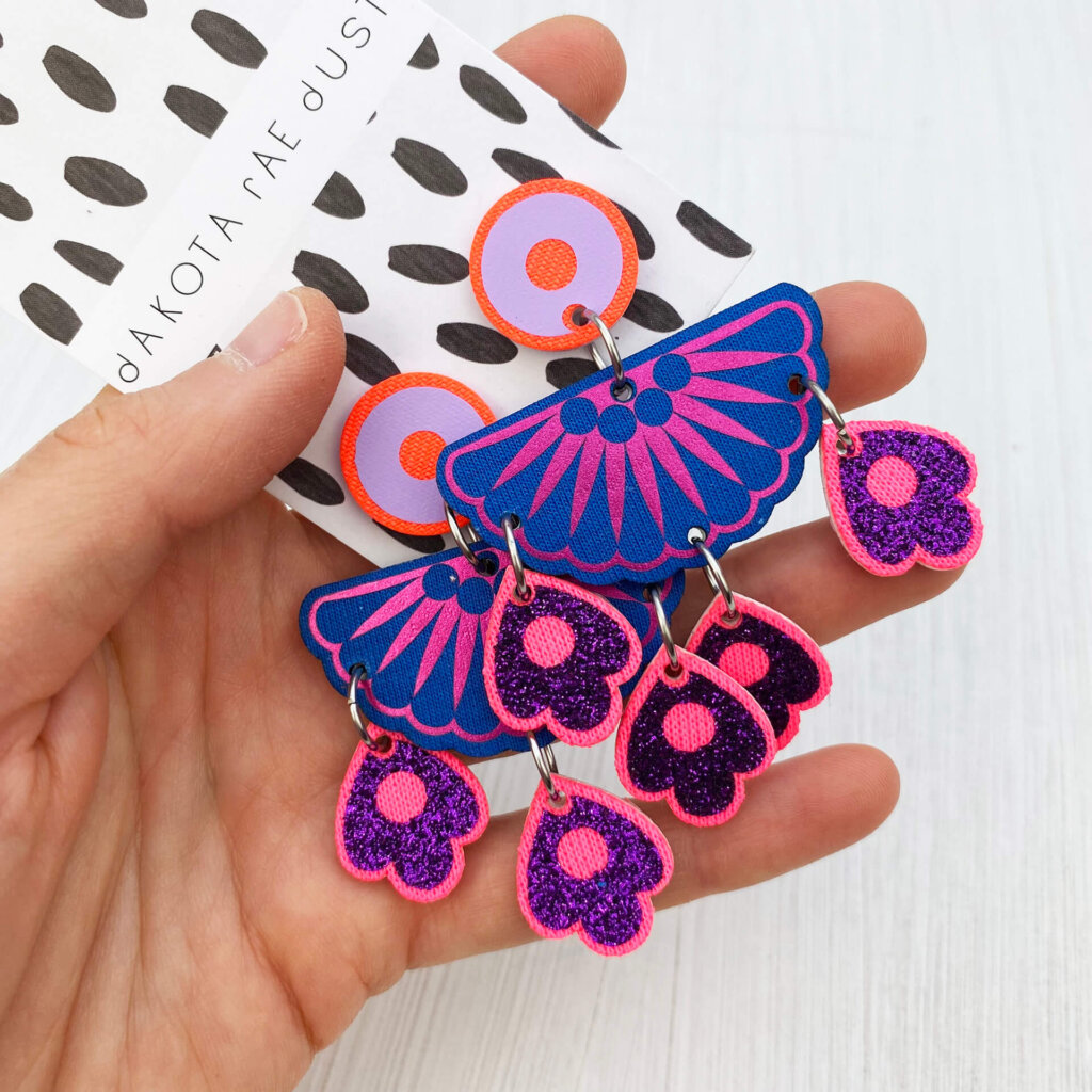 A pair of bold blue and pink jangly droplet earrings mounted on an a black and white patterned dakota rae dust branded card are lying on an open hand on an off white background
