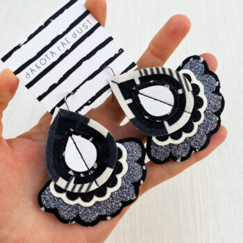 A pair of monochrome fabric oversize earrings in black and white mounted on a black and white patterned, dakota rae dust branded card are held in an open hand