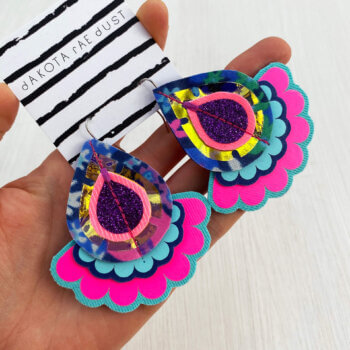 A pair of oversize floral fabric earrings mounted on a black and white patterned, dakota rae dust branded card are held in an open hand