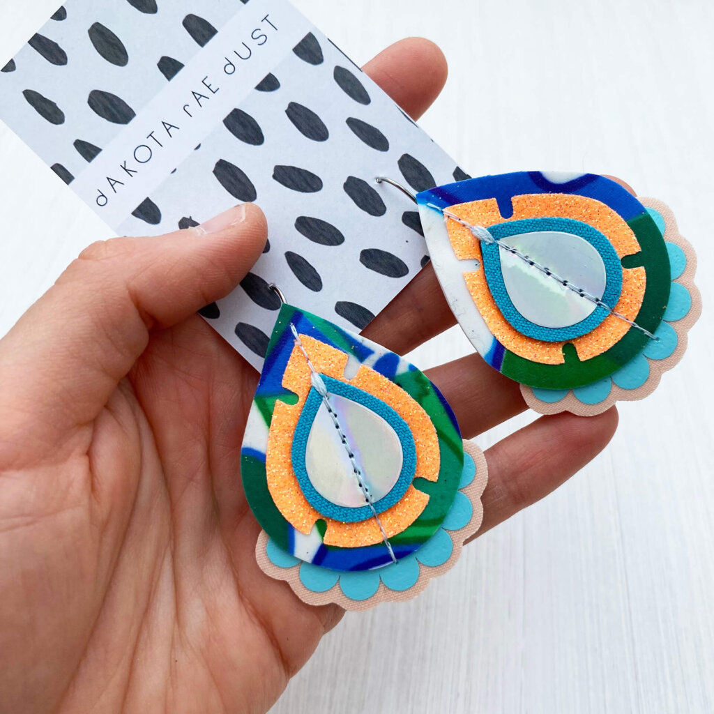 A pair of peach, green and blue recycled fabric earrings held in a open hand